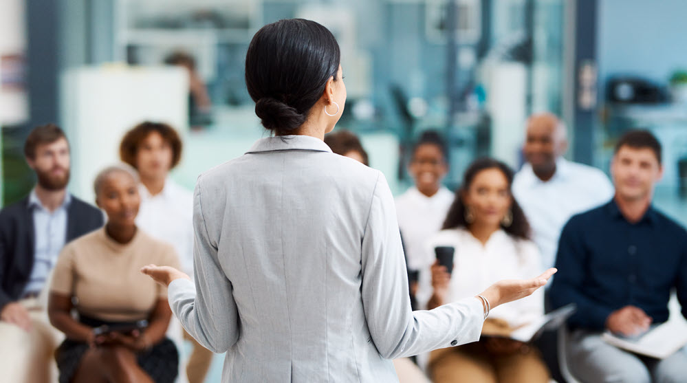 A close up of someone sitting at a desk and writing something. They appear to be in a meeting with other associates.