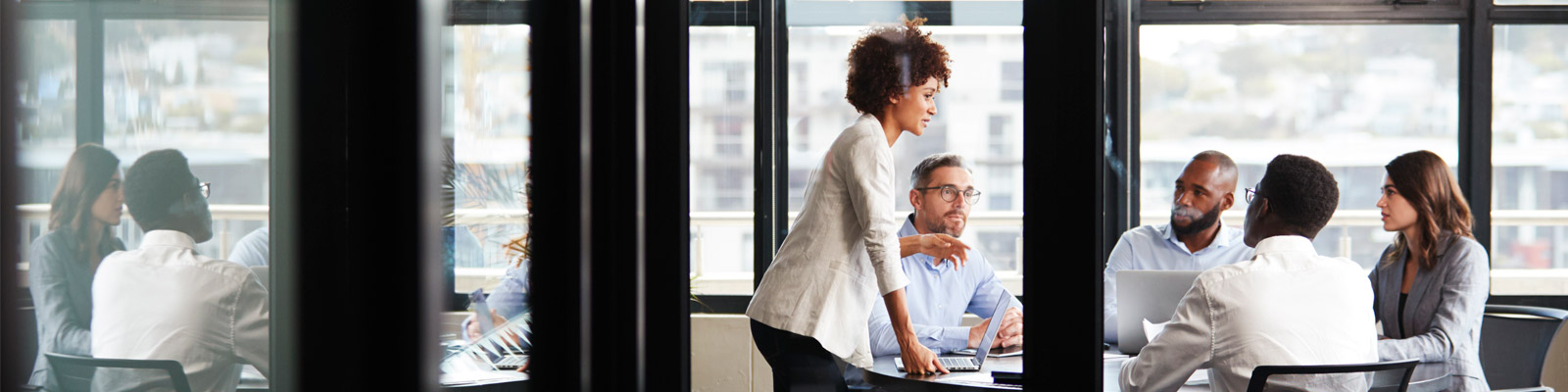 A businesswoman stands addressing colleagues at a meeting, seen through glass wall.