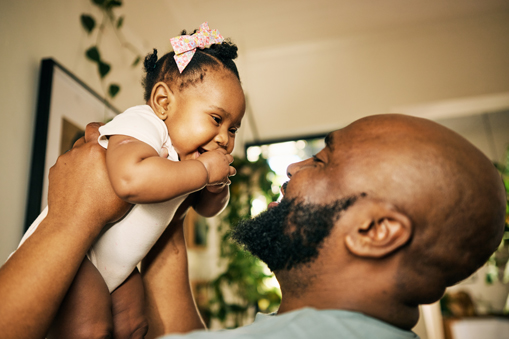 Cheerful baby girl giggles while being held up in the air by her happy father